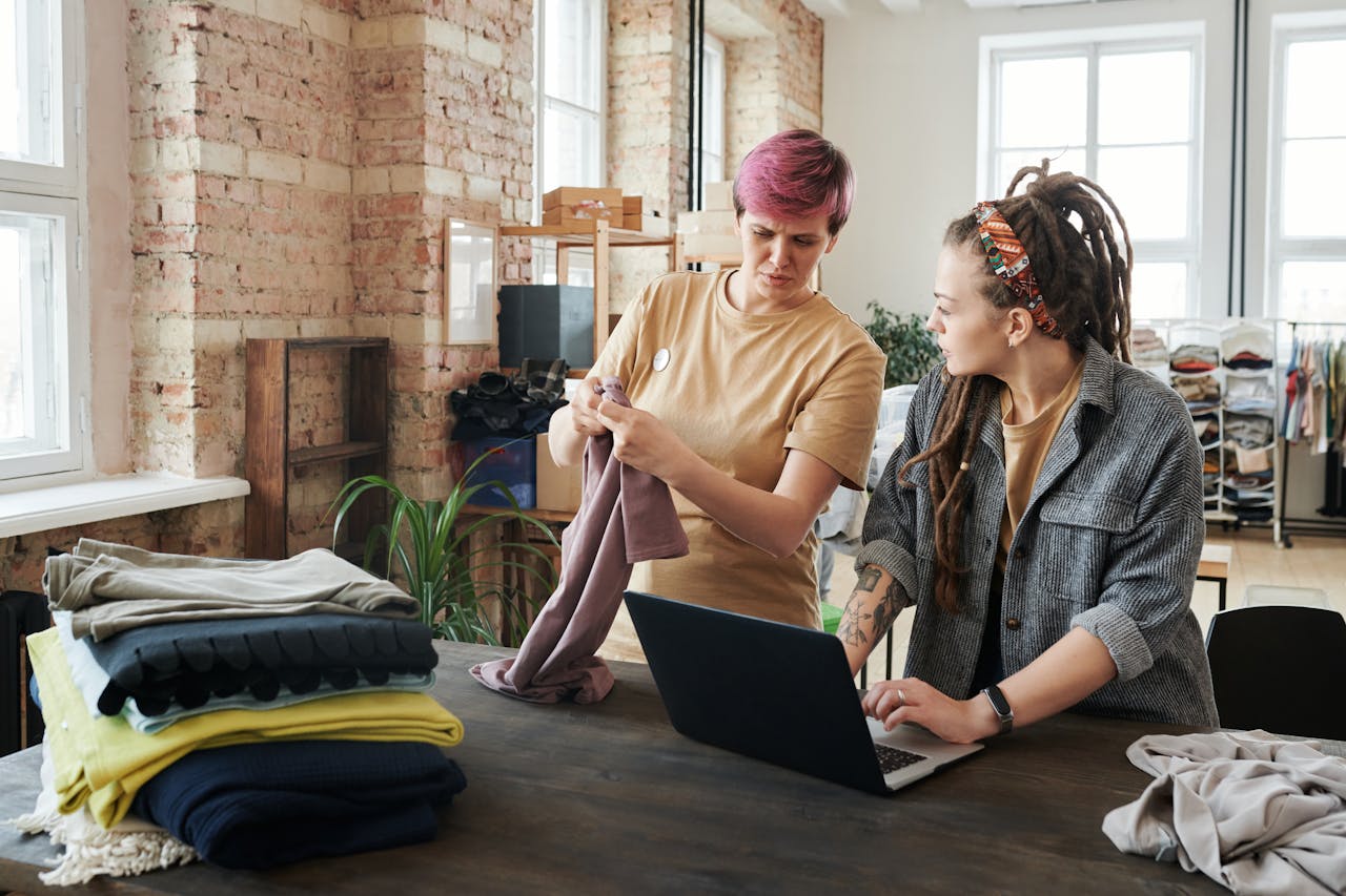 Two women sorting clothing donations using a laptop in a bright urban setting.